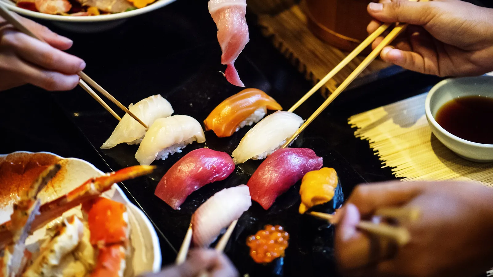 Sushis et makis présentés sur un plateau noir, baguettes en main autour de la table